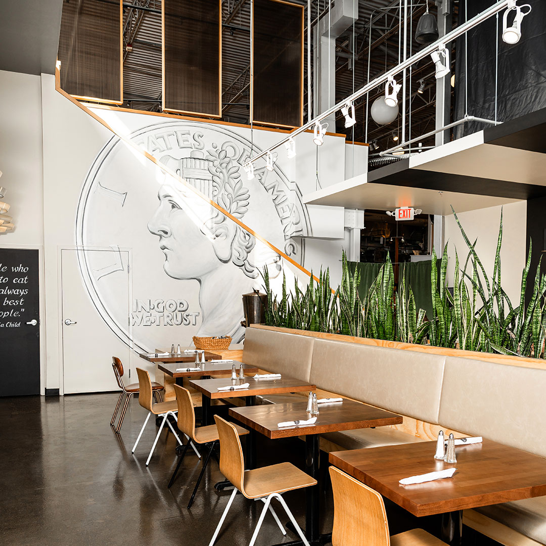 Moody view of Dime Store Rochester Hills dining room with mural, settee seating, and large planter