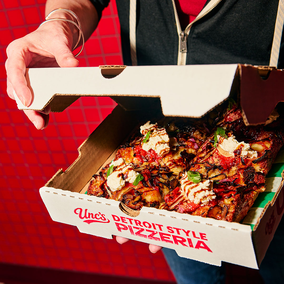 Person holding a slightly open Unc’s Pizza box with a pizza inside.
